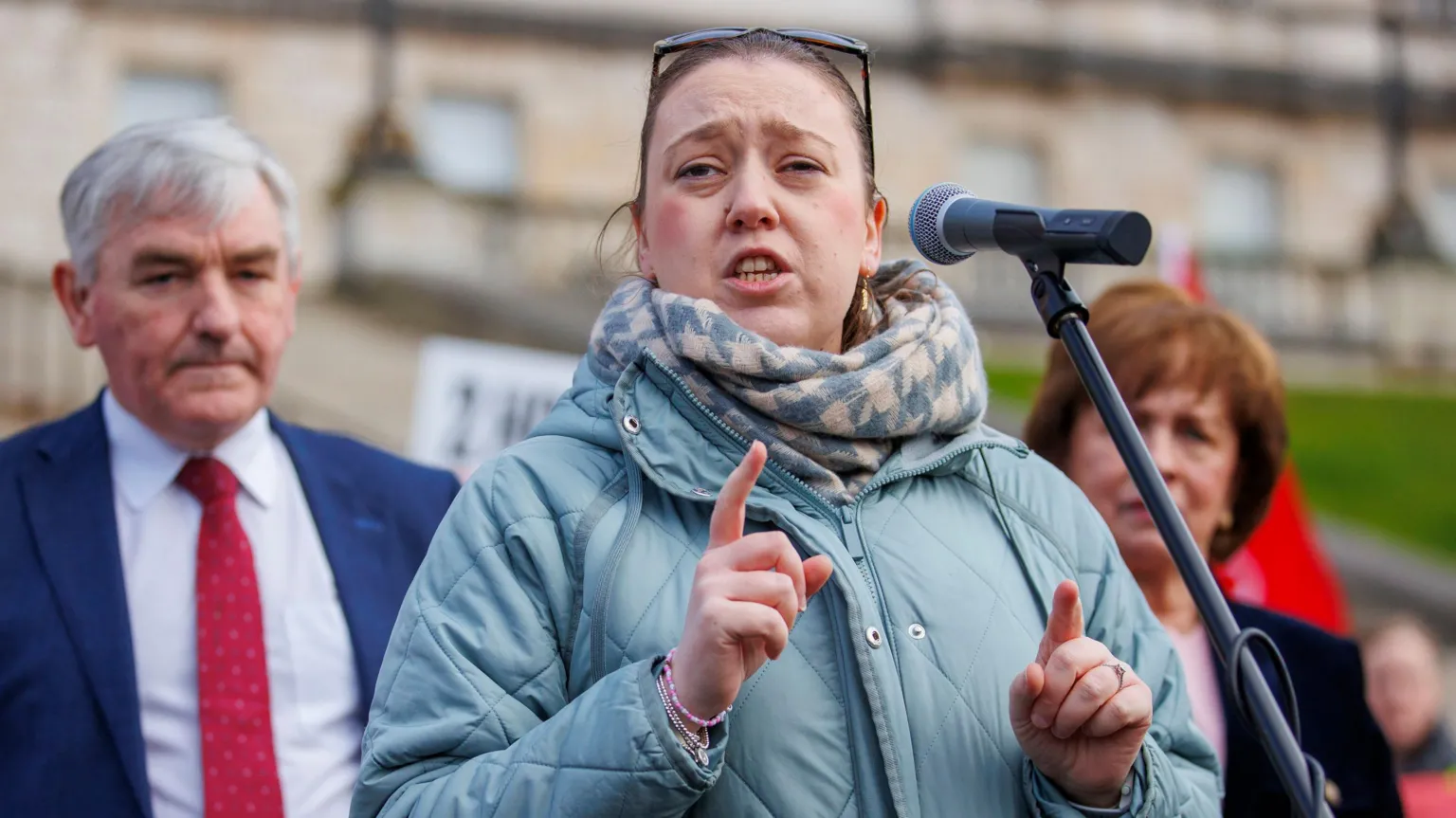 DUP MLA Deborah Erskine, standing in front of a microphone, wearing a light blue coat and a blue and cream scarf. There is a man standing behind her right shoulder. He has grey hair and is wearing a white shirt, blue suit and red tie. There is a woman behind her left shoulder, with short brown hair. She is wearing a pink top and a black suit.