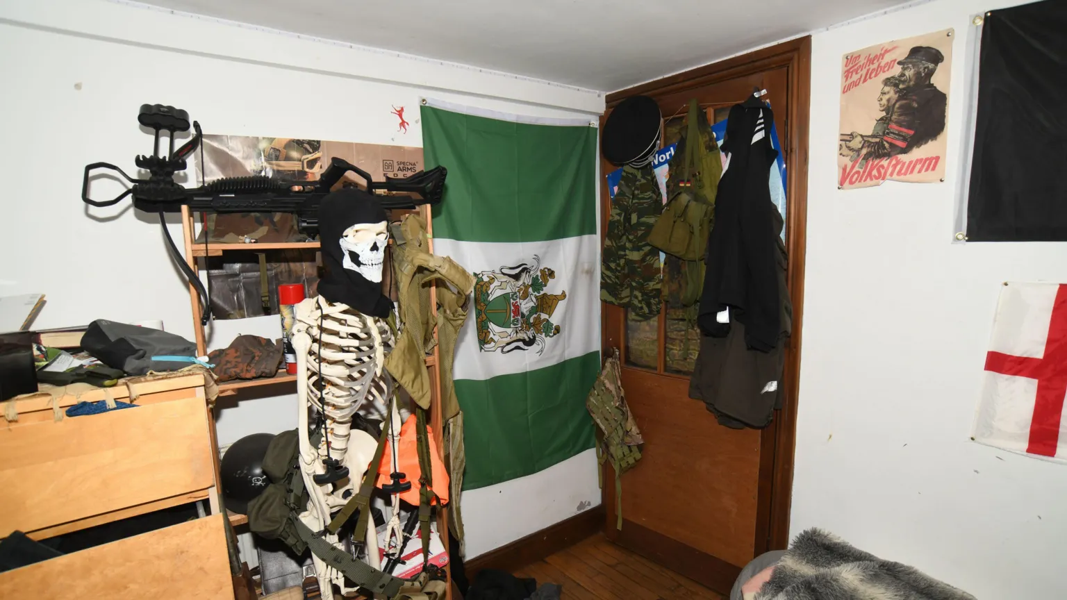 Counter Terror Policing North East A view of the corner of the accused's bedroom. Military clothing is hanging on the back of the door, a poster with historical Nazi propaganda on it hangs on the wall and a large weapon and plastic skeleton wearing a mask are in the foreground. There is also a green and white flag hung sideways between the door and the skeleton.