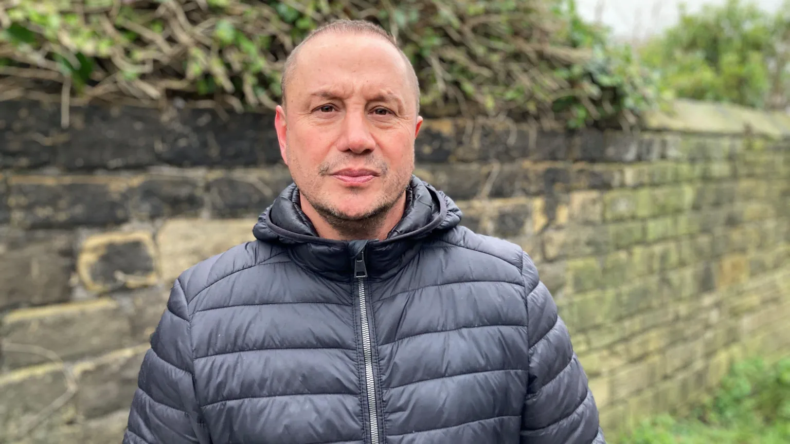 A man with short hair, wearing a blue/grey puffer jacket, looks straight at the camera, with a stone wall behind him.