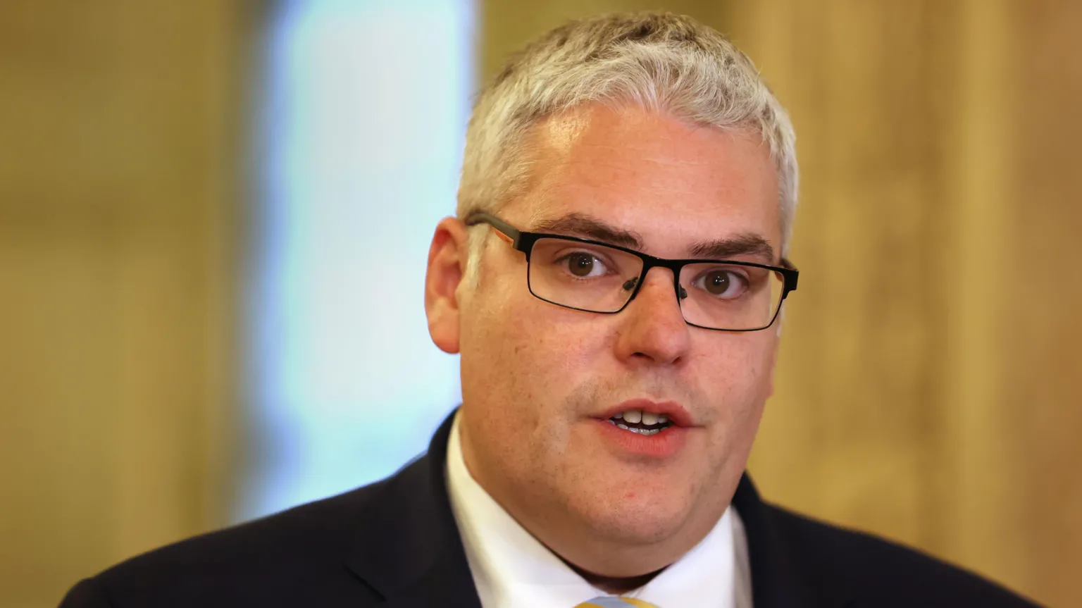 Gavin Robinson is speaking in the Great Hall at Stormont. He has grey hair and is wearing glasses and a suit. 