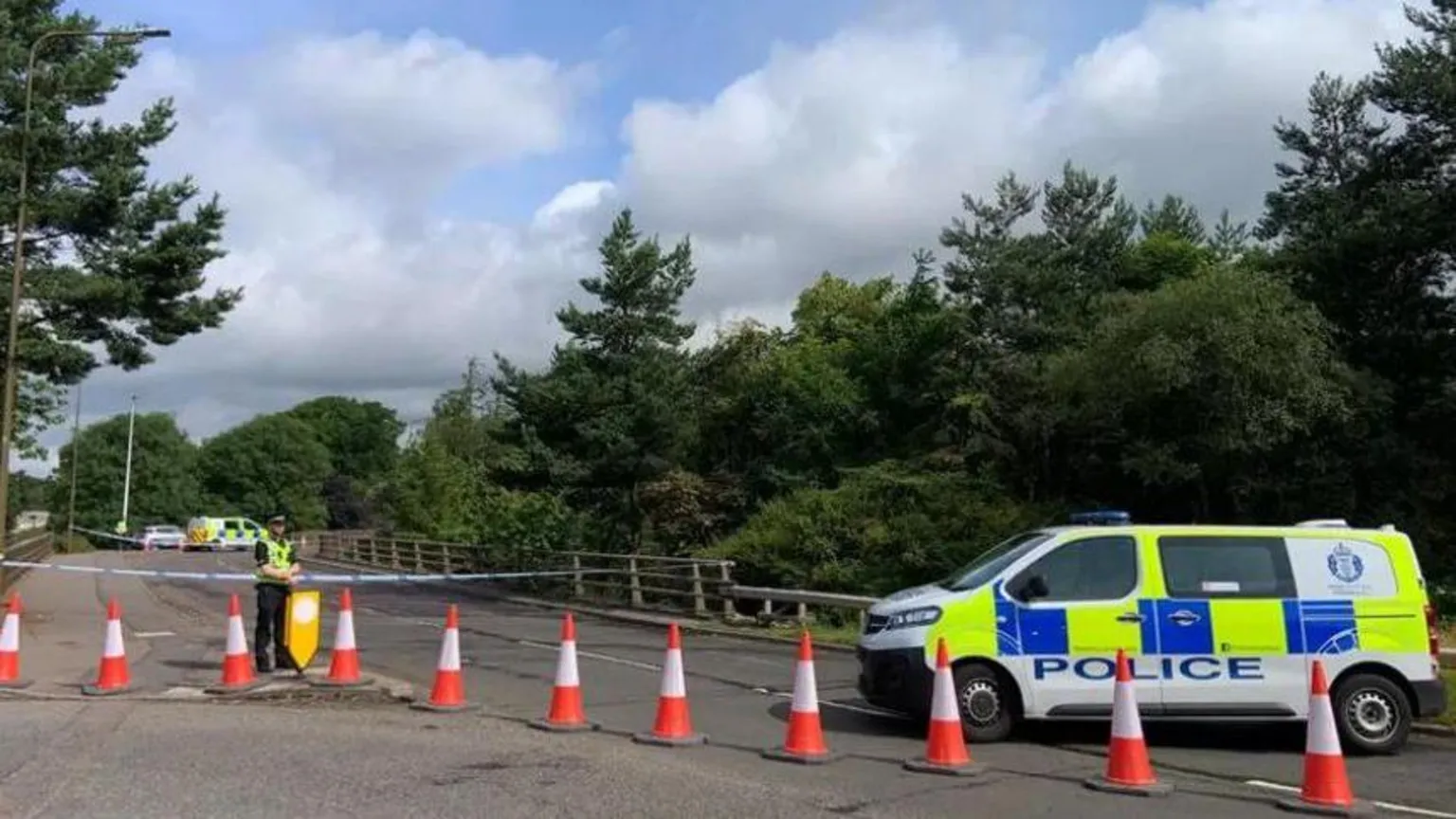 a police van parked behind some orange traffic cones, in front of a police cordon being guarded by a police officer.