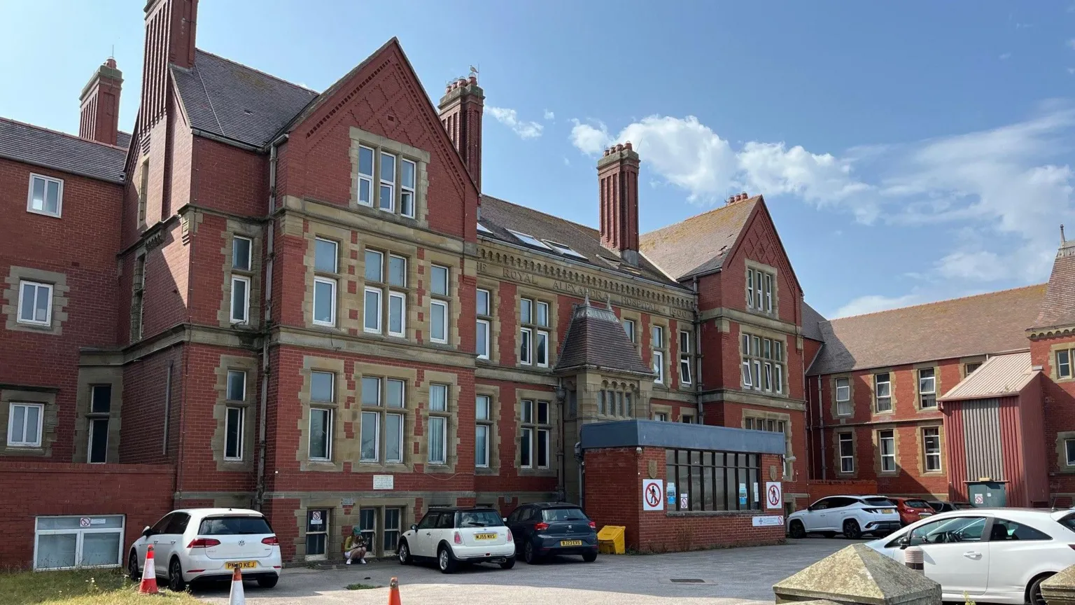 The Royal Alexandra Hospital in Rhyl, Denbighshire, with blue sky behind it. It is a large red brick building with four horizontal rows of windows and a few cars parked in front.