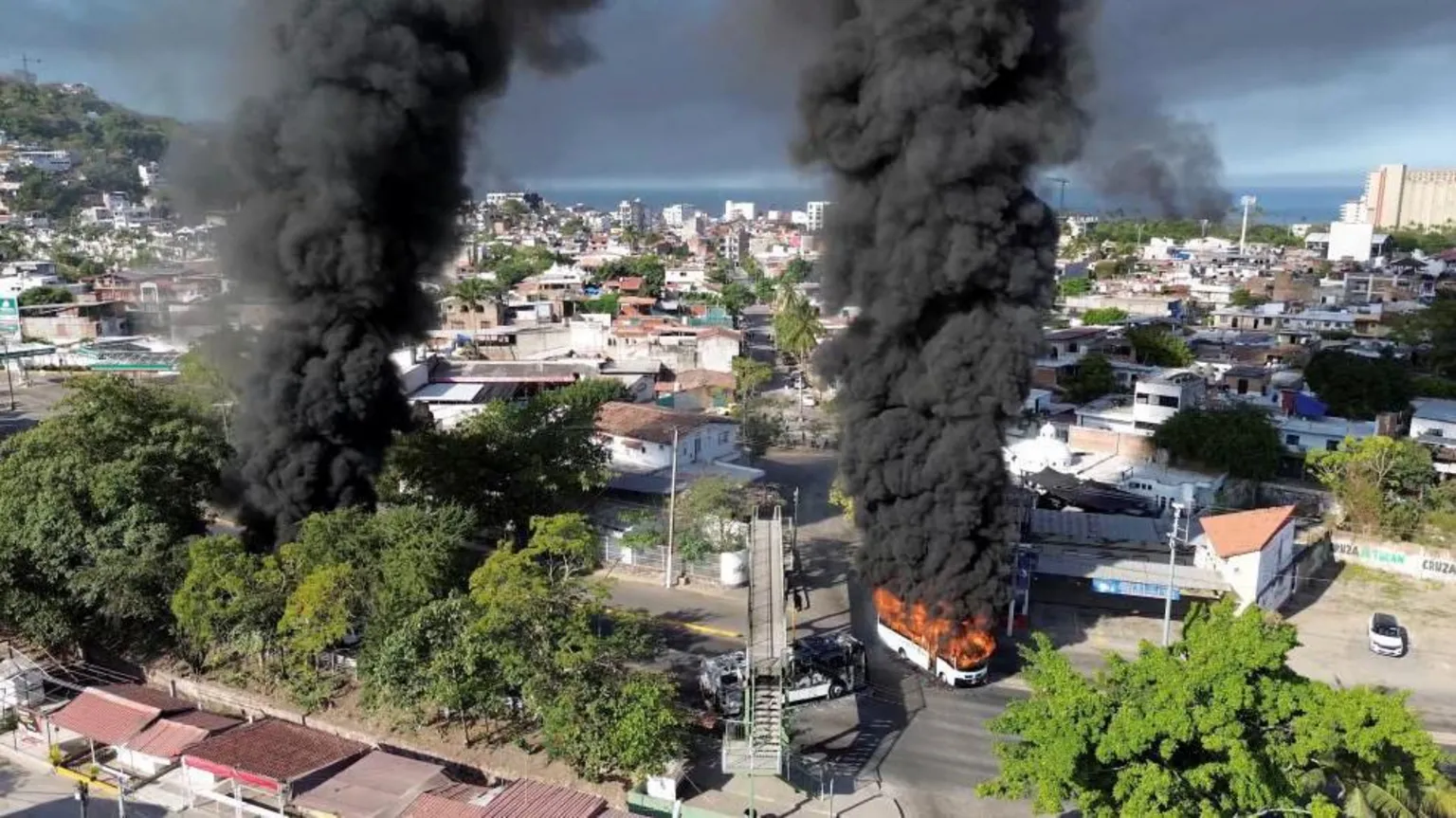MoreLifeDiaries via Screengrab from a drone video showing black smoke rising in the sky from several locations, including from a bus that has been set on fire. The smoke is rising between buildings and greenery. 