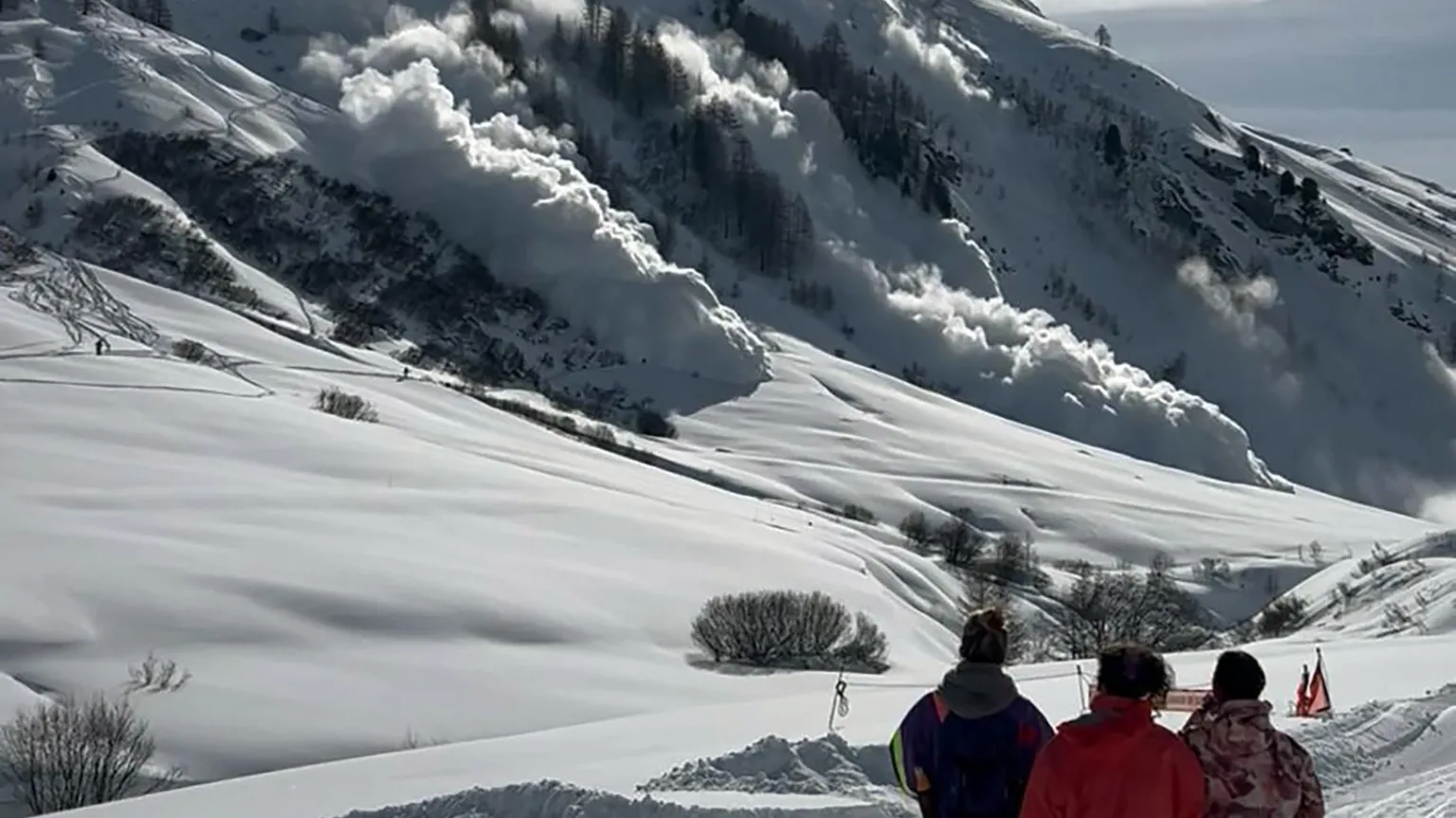 Antoine Martignon Three people in foreground watch as an avalanche descends a snowy mountainside from left to right at Val d'Isere