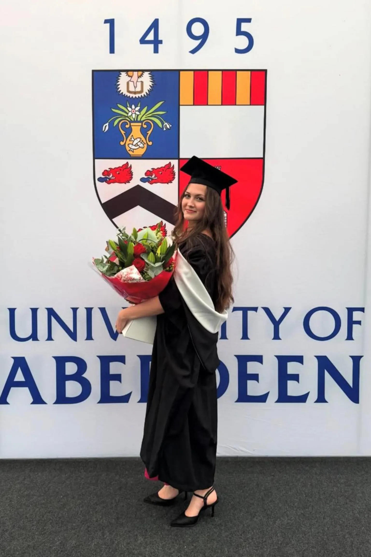 Viktoriia Sirenko Anastasiia, dressed in the graduation robe and hat, holding a bouquet of flower is standing in front of the coat of arms and logo of the university of Aberdeen