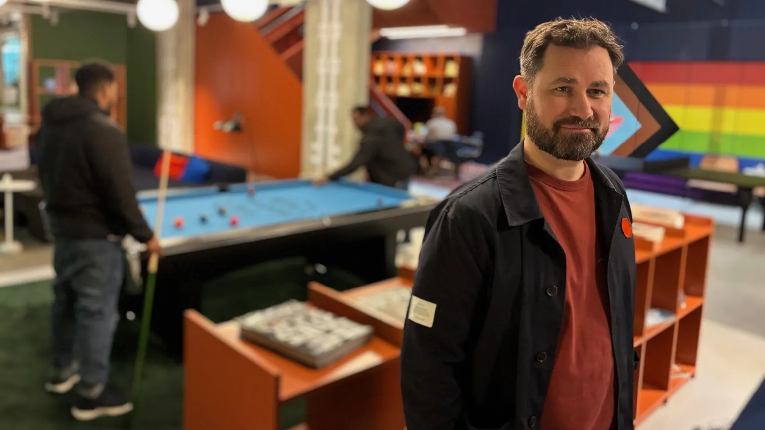 A man with short brown hair and a bear is wearing a black jacket over a red top. He is standing in the lobby with two people playing pool behind him