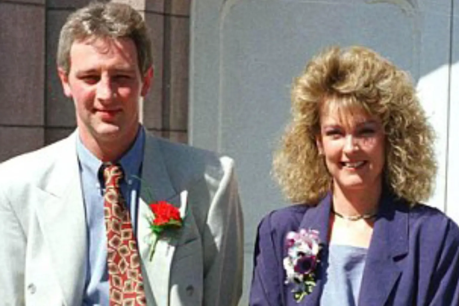 Nat and Arlene Fraser dressed for a wedding. She is on the right, with light brown permed hair and is wearing a purple jacket with a flower in the buttonhole. He has short, light brown hair and is wearing a light coloured suit, blue shirt and a patterned tie. He has a red rose in his buttonhole.