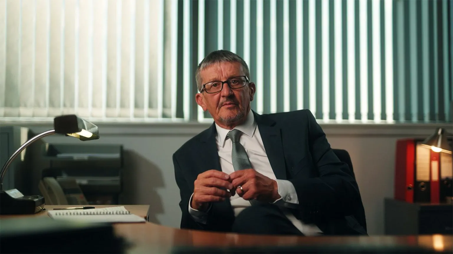 Sean De Francesco/Firecrest Films Alan Smith sitting behind a desk, with a lamp to his left, in front of a window with vertical blinds. He is wearing glasses and is dressed in a black suit with a white shirt and a green tie.