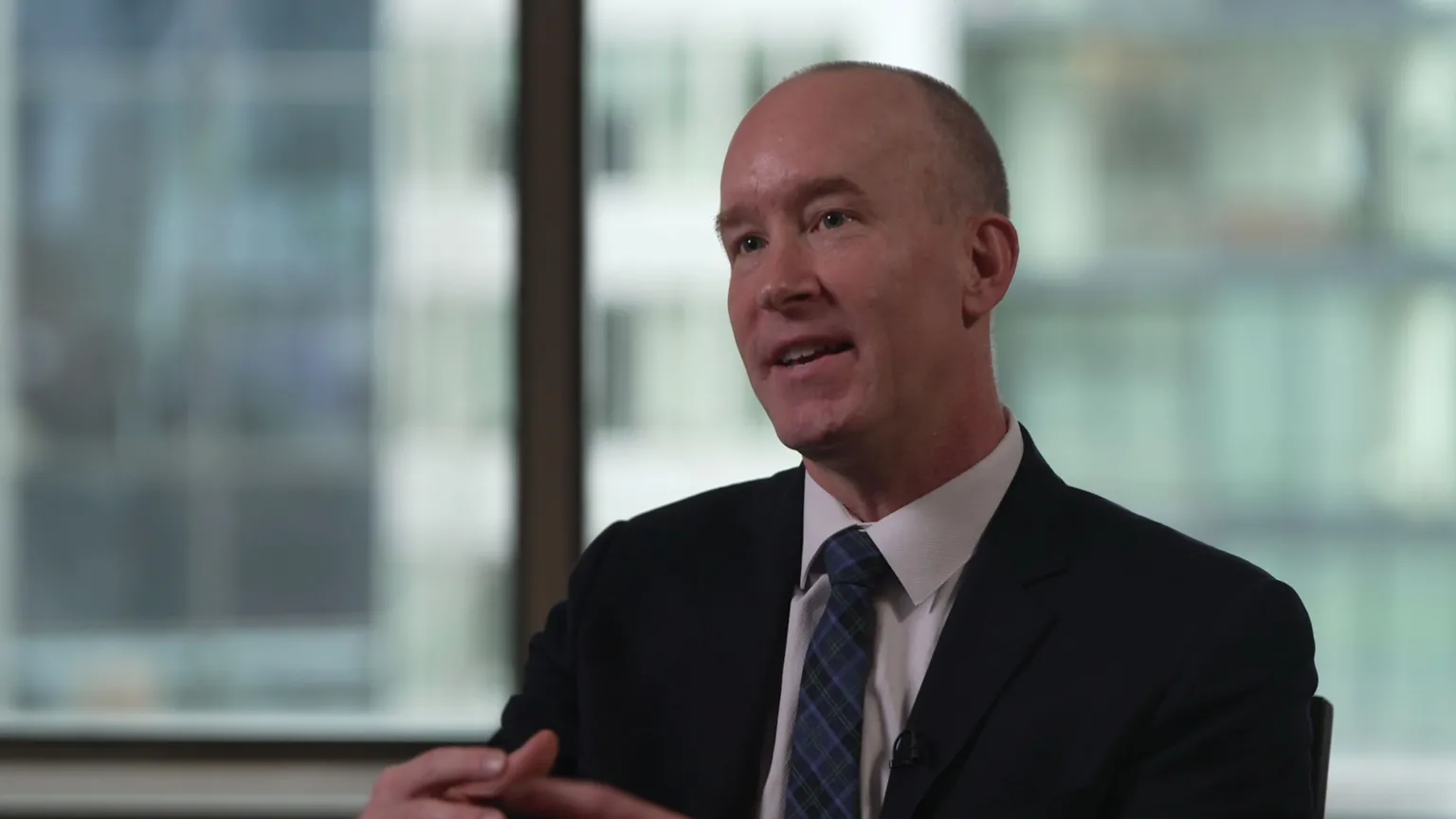 A man in a dark suit looks to the left of the camera, with his hands clasping in motion. He is wearing a white shirt and blue and black chequered tie. The man has short grey hair and is sitting in an office, with the windows in the background out of focus. 