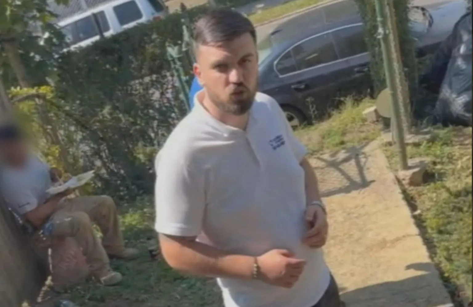 Ginia Hines A man with short dark hair and a neatly trimmed beard is wearing a white t-shirt. He is standing on a small pathway in the front garden of a home, with another man in work clothing sitting behind him eating. The second man's face is blurred. Cars, a hedge and a small patch of grass are visible in the background. 