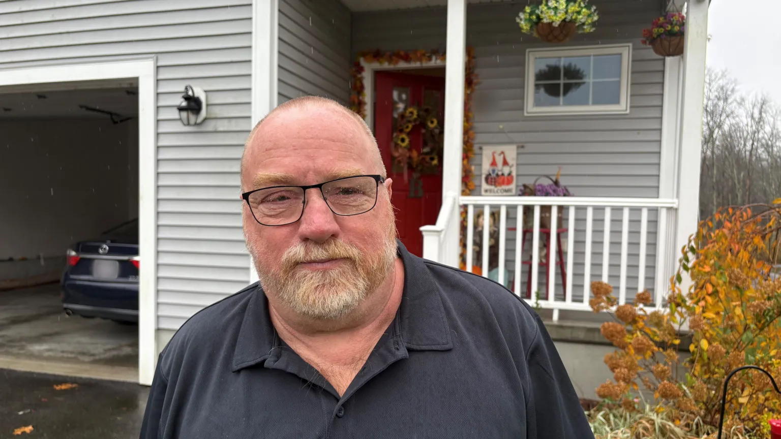 A balding man in a navy polo neck t-shirt stands outside his home, which is made of wooden grey slat and has a small white painted porch and garage open. He has a short beard which is turning grey and is wearing glasses which have specks of rain visible. 