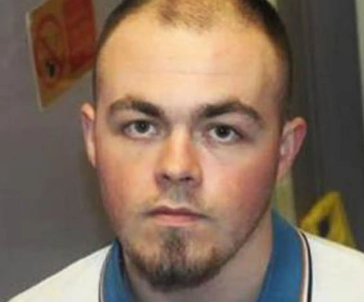 Department of Justice Northern Ireland A man in a white t-shirt with a blue polo neck collar is looking directly at the camera. He has dark hair and brown eyes. The man has a short goatee style beard. The background shows some health and safety signage. The rest of the image is blurred. 
