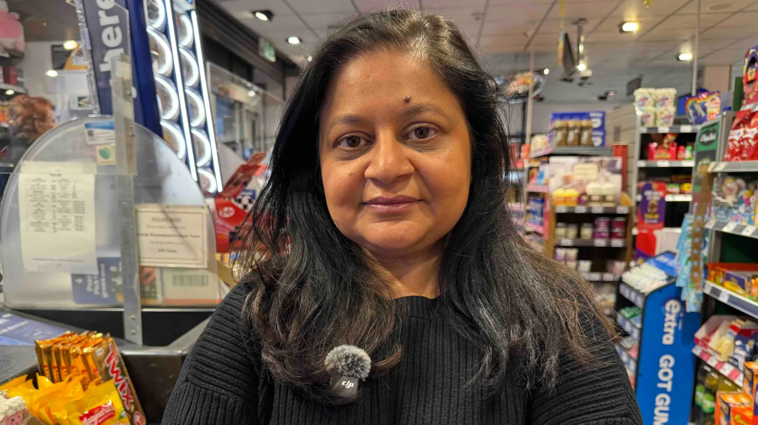 Sunita is a shopkeeper in Sheffield. She is standing to the right of her shop counter. Lottery scratchcards are on sale on the left on the counter, behind Sunita are rows of aisles in her convenience store, and to the right we can see the ends of aisles with promotional goods. Sunita is in her 50s, has long black hair past her shoulders and is wearing a black jumper.