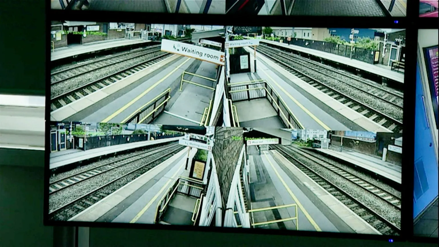 A surveillance screen shows six CCTV angles of Sandwell & Dudley station. Each feed captures empty platforms, railway tracks, yellow safety lines, and signs for the waiting room. The station appears clean and quiet with no passengers present.