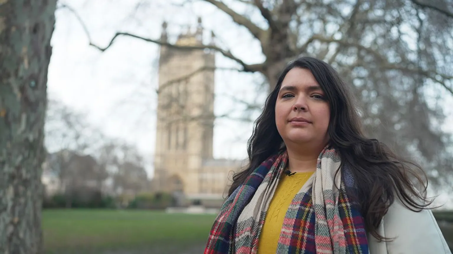 Rebecca stands in a park with leafless trees, facing the camera. She wears a colourful plaid scarf, a yellow shirt, and a light-coloured coat with a small microphone clipped to it. Behind her is the Palace of Westminster, including the Victoria Tower, under an overcast sky.