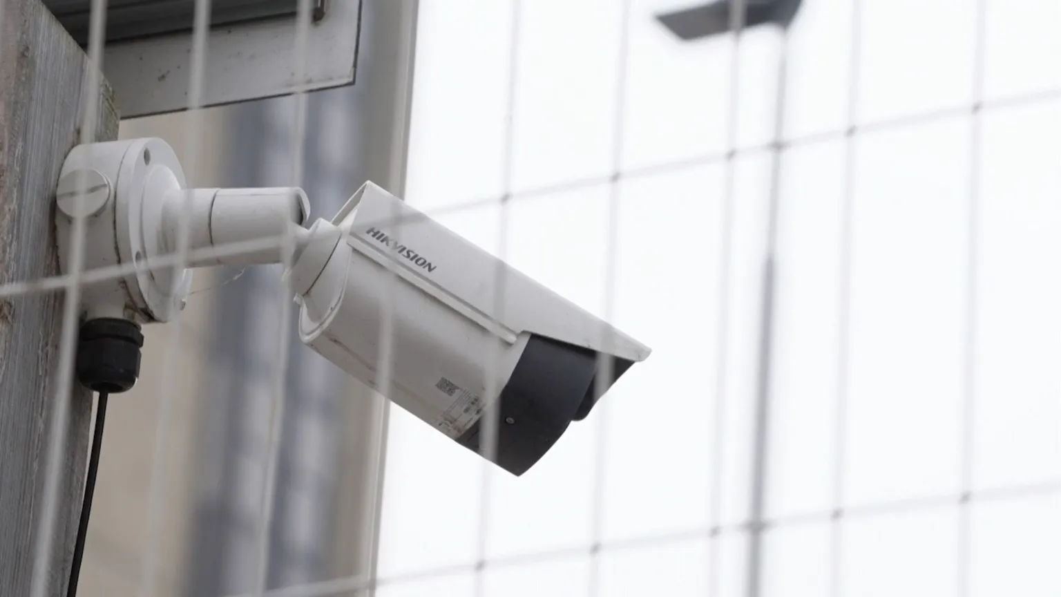 A white CCTV camera is mounted on a wooden pole, angled downward as if monitoring an area. A black cable runs from the camera, and the background shows a grid‑style fence and out‑of‑focus architectural structures.