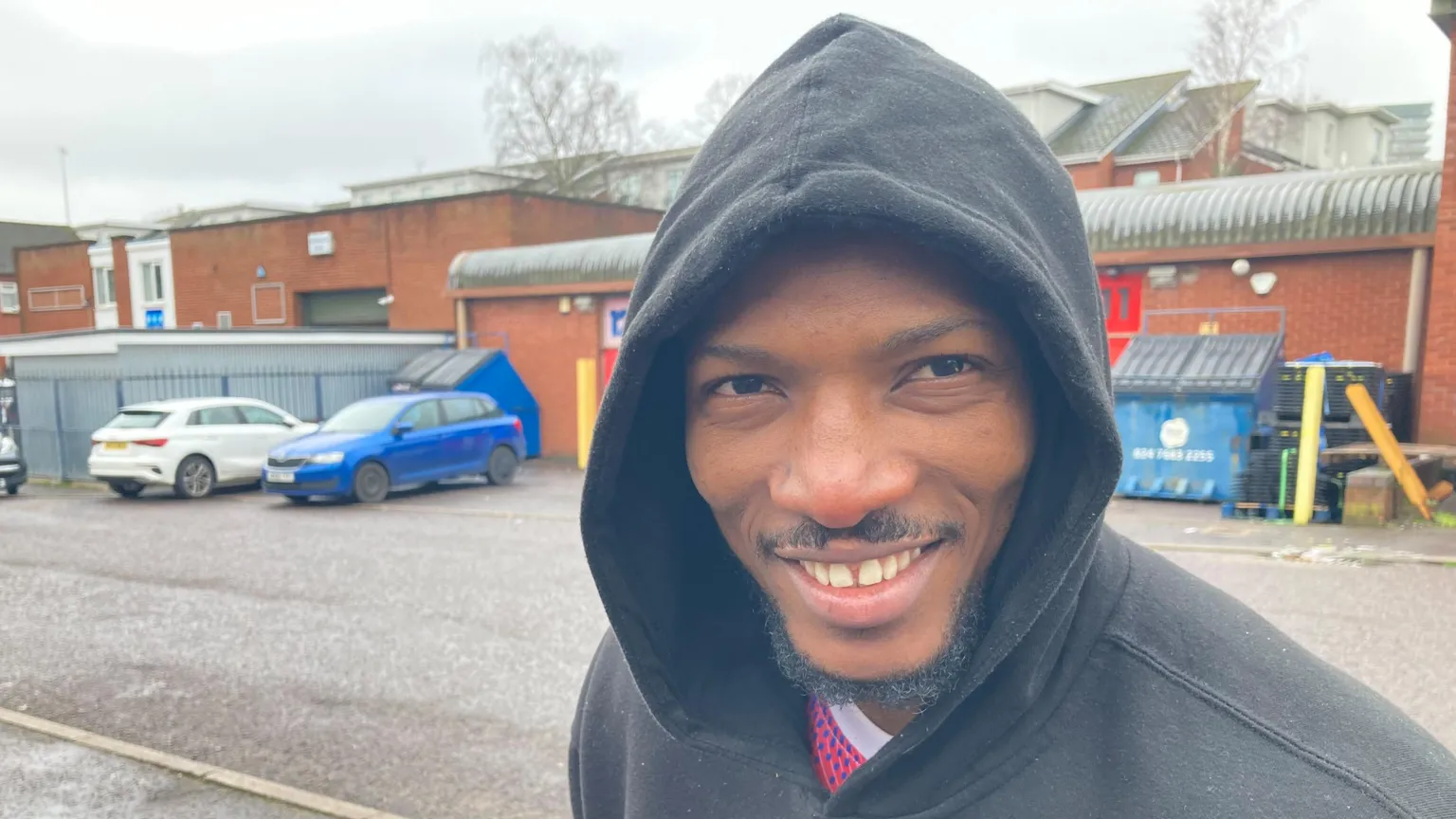 Mohammed is wearing a black hoodie over a pink-and black spotted sports top, with a white collar. He is standing outside the Clothing Coventry warehouse and smiling at the camera.