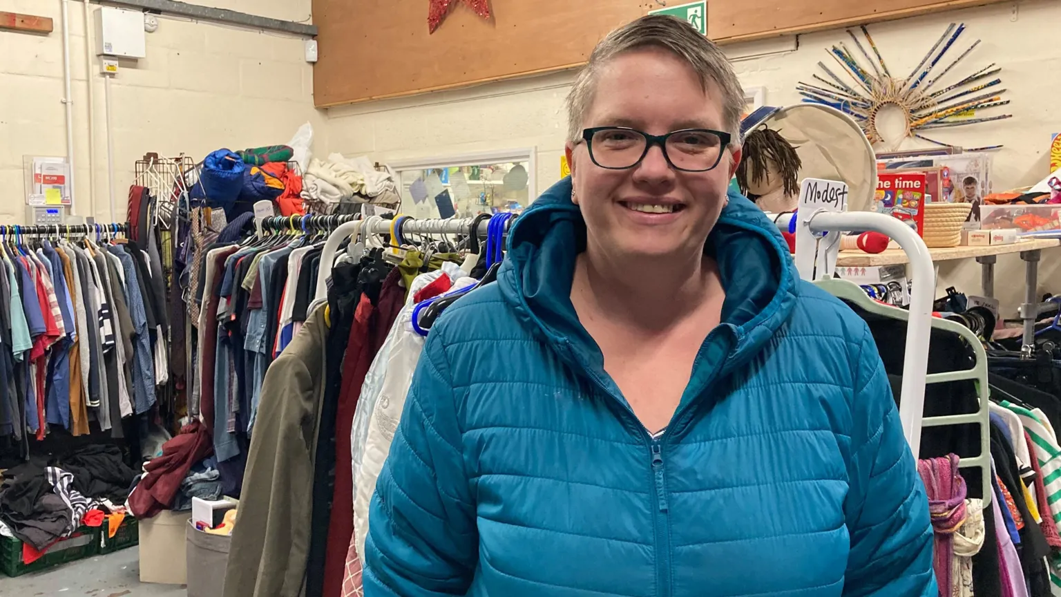 Katie Wilson is wearing a blue hooded jacked and standing in front of two racks of clothing at the Clothing Coventry warehouse. She has a pair of black-framed glasses and has short blonde hair.