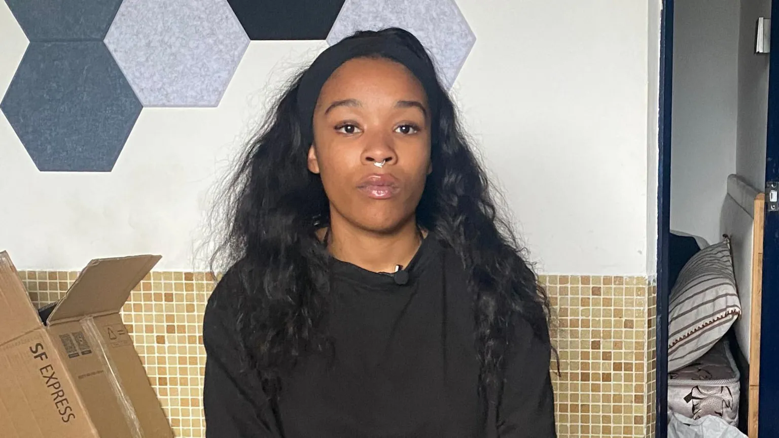 Isabel Rose is seen wearing a black top and a small nose ring, sits facing the camera indoors. A cardboard box is visible beside her and hexagonal acoustic panels are mounted on the wall behind her.