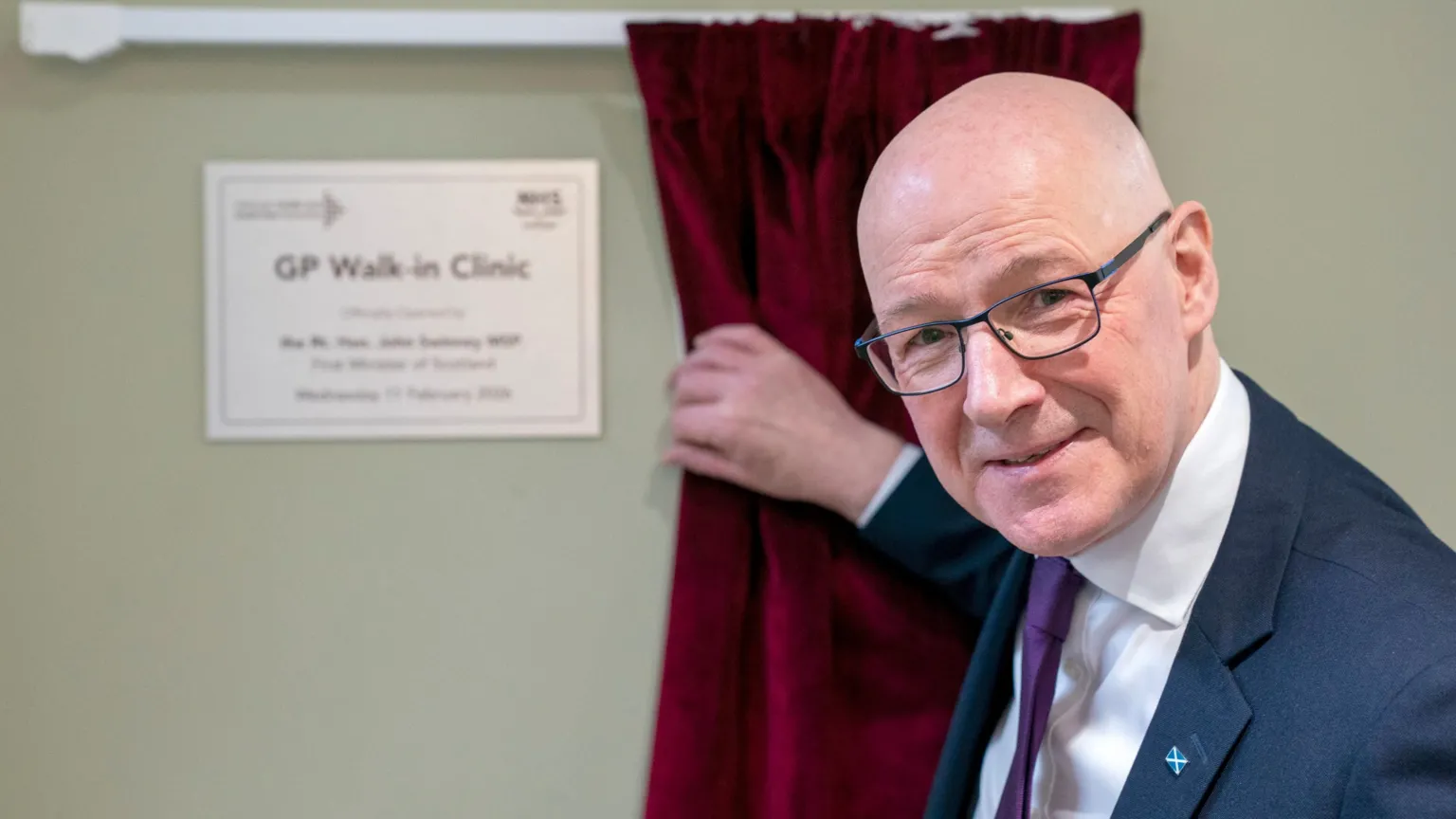  First Minister John Swinney, a bald man with dark framed glasses wearing a navy blue suit jacket with a white shirt and purple tie, unveils a sign reading GP Walk-in Clinic. His hand on on a red velvet curtain on a white rail. His opening the first GP walk-in clinic at Wester Hailes Healthy Living Centre in Edinburgh.