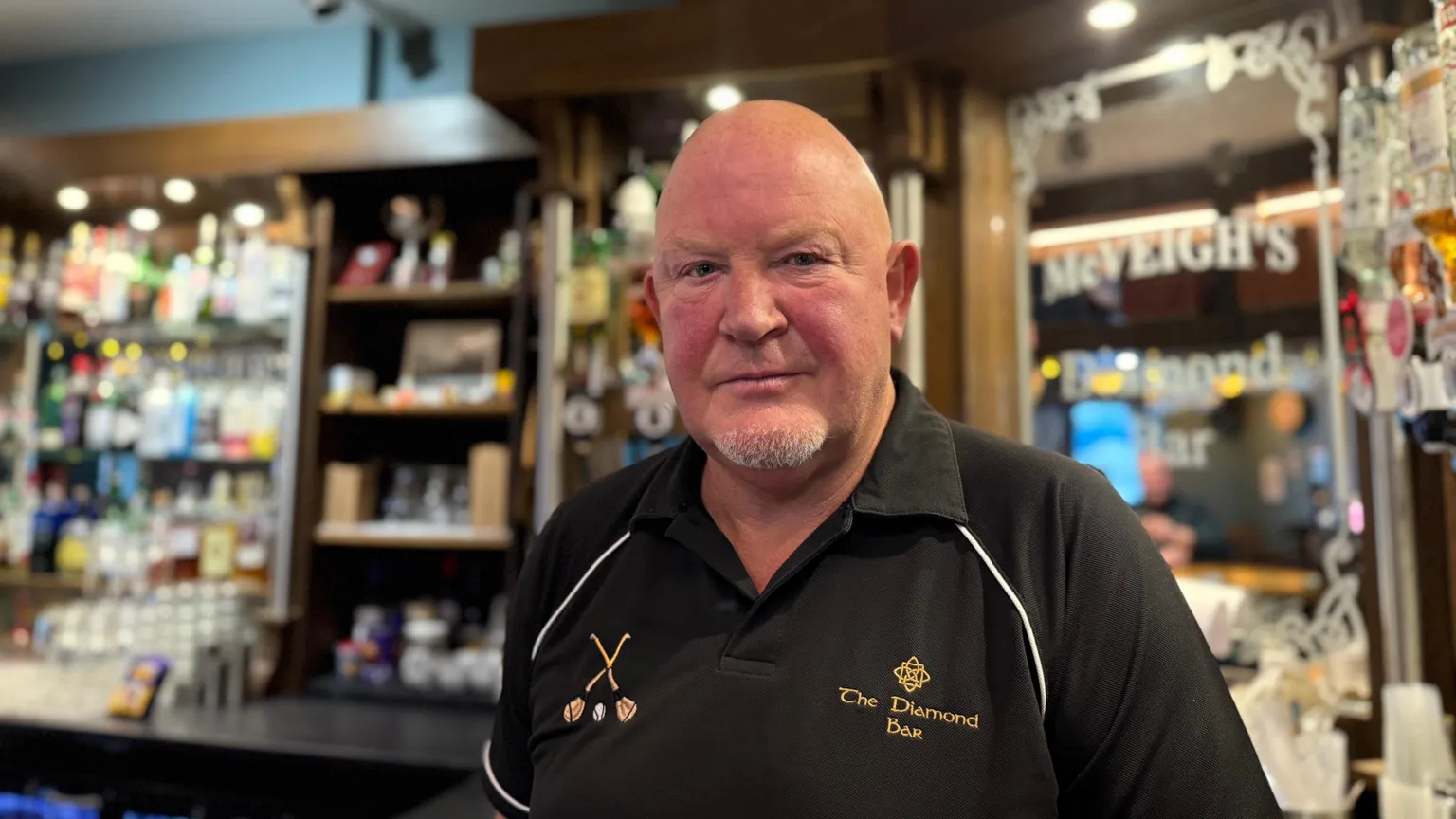 Charlie McVeigh is an older, bald man who is looking at the camera with a neutral expression. He is wearing a black, polo shirt with The Diamond Bar embrodiered on the chest. He is standing in front of a stocked bar, but the background is blurred.