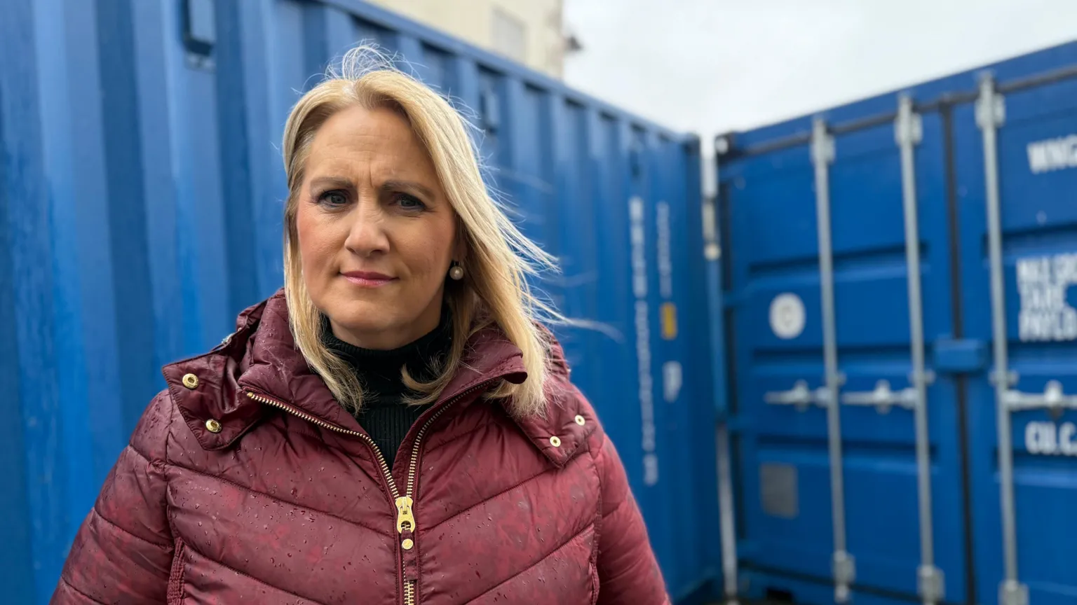 Claire Parker stands in front of blue shipping containers and is looking at the camera with a neutral expression on her face. She has shoulder-length blonde hair and is wearing a zip-up burgundy jacket.