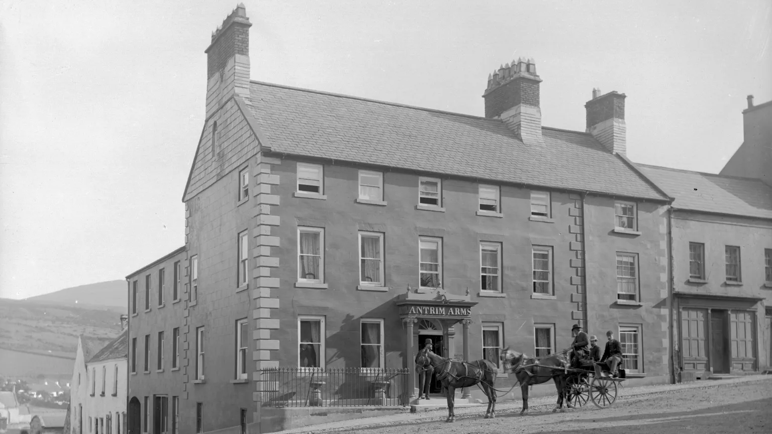 Courtesy of National Museums NI A black and white archive photo of the Antrim Arms hotel in Ballycastle. The hotel is a large Georgian-era building on a corner site. There is a cart pulled by two horses waiting outside the entrance. Three well-dressed men are sitting on the cart. 