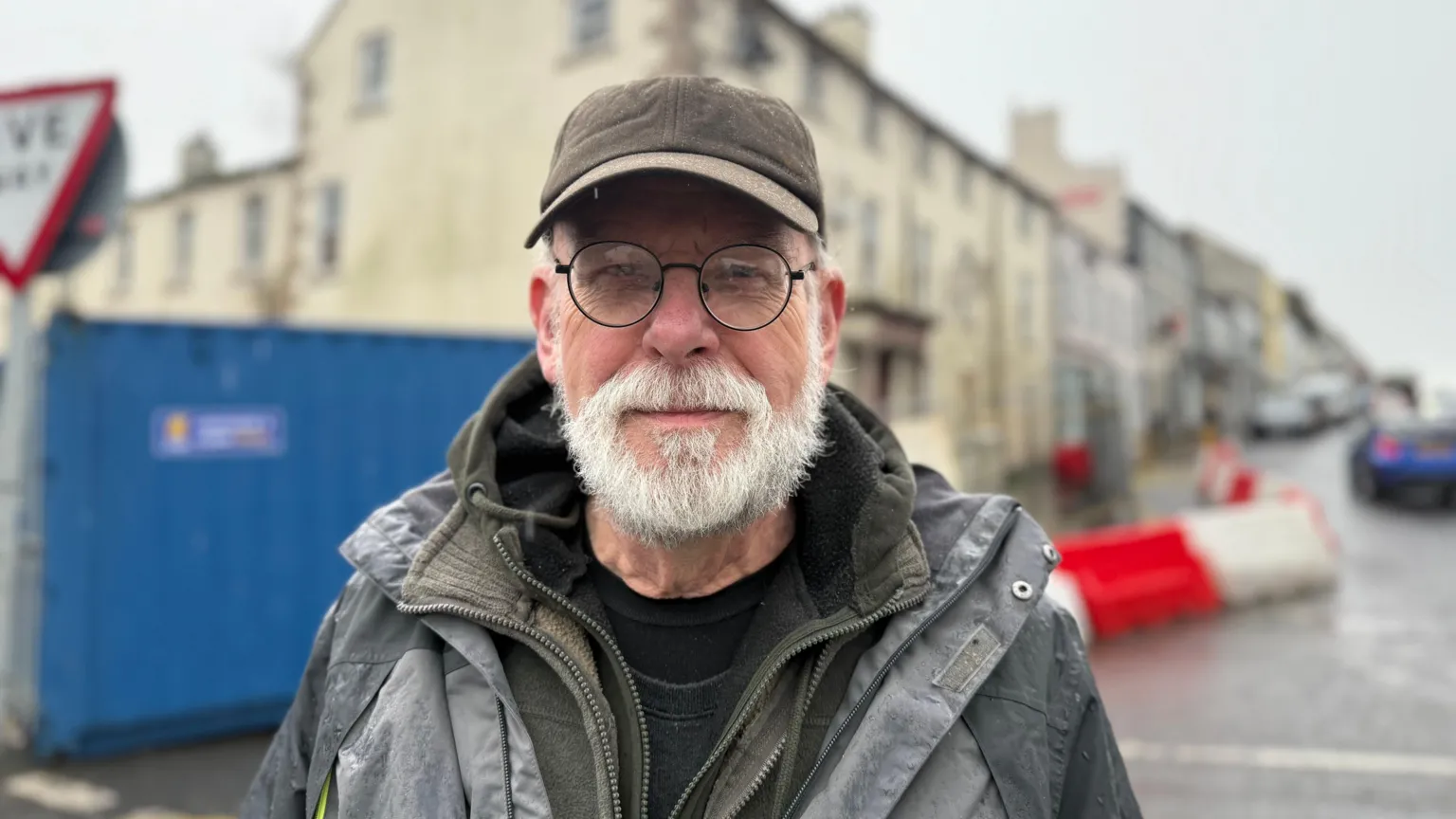 John Anderson is an older man with a long white beard and circular glasses. He is wearing a grey hat, a rain coat and a green zip up jumper. He is standing just in front of the disused building but the background is blurred.