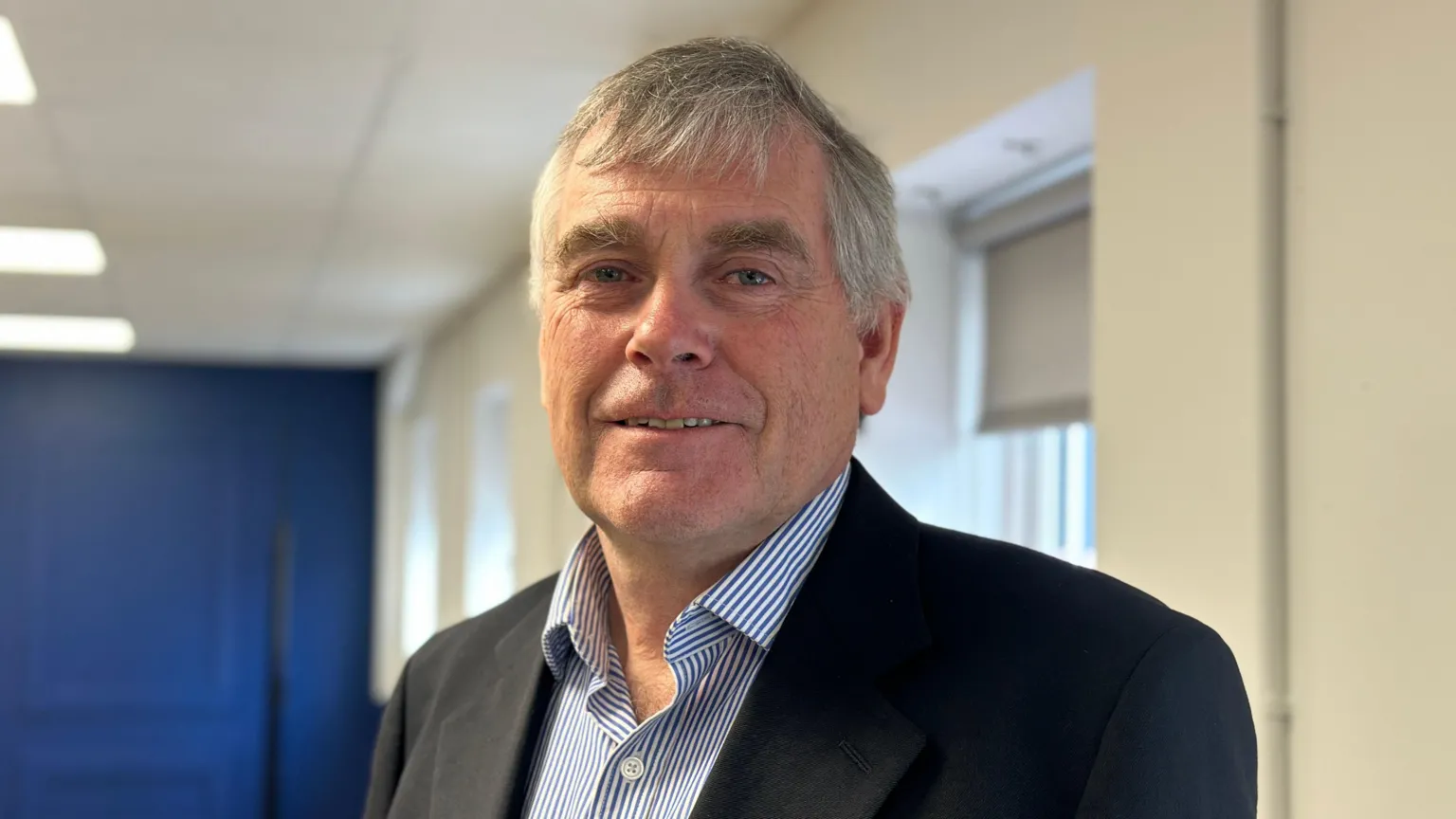 Declan Wright is an older man with short, grey hair. He is smiling at the camera. He is wearing a black suit jacket and a blue and white striped shirt.