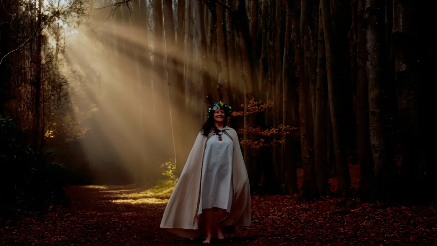 Cherry Tree Deborah Rose Hālani stands barefoot in a wooded area wearing white robes. Rays of sunlight shine through the cracks in the trees illuminating the leafy ground underfoot.