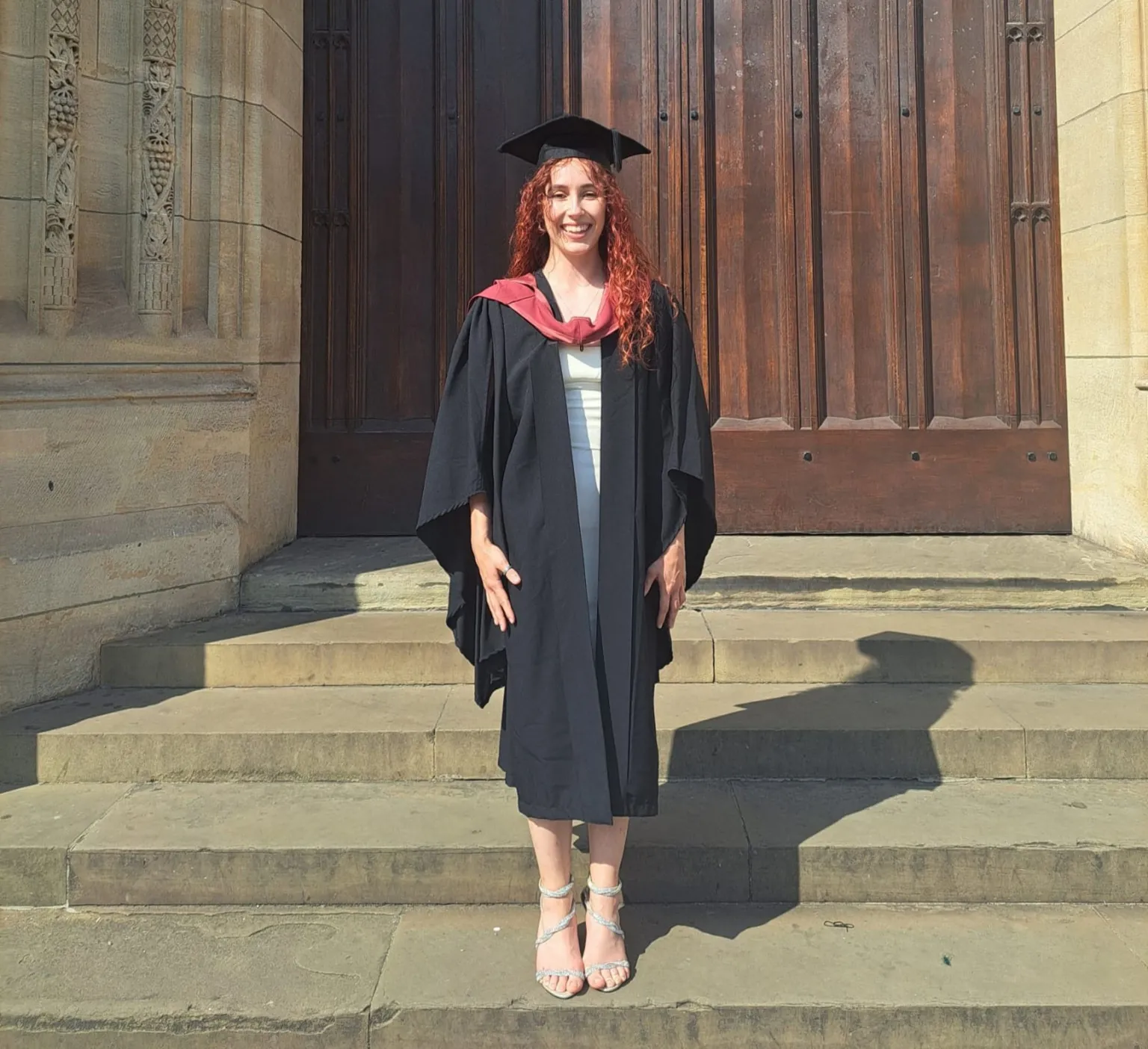 Caitlin stands outside a university building on the day of her graduation. She is wearing a graduation cap and gown and has long, red hair.