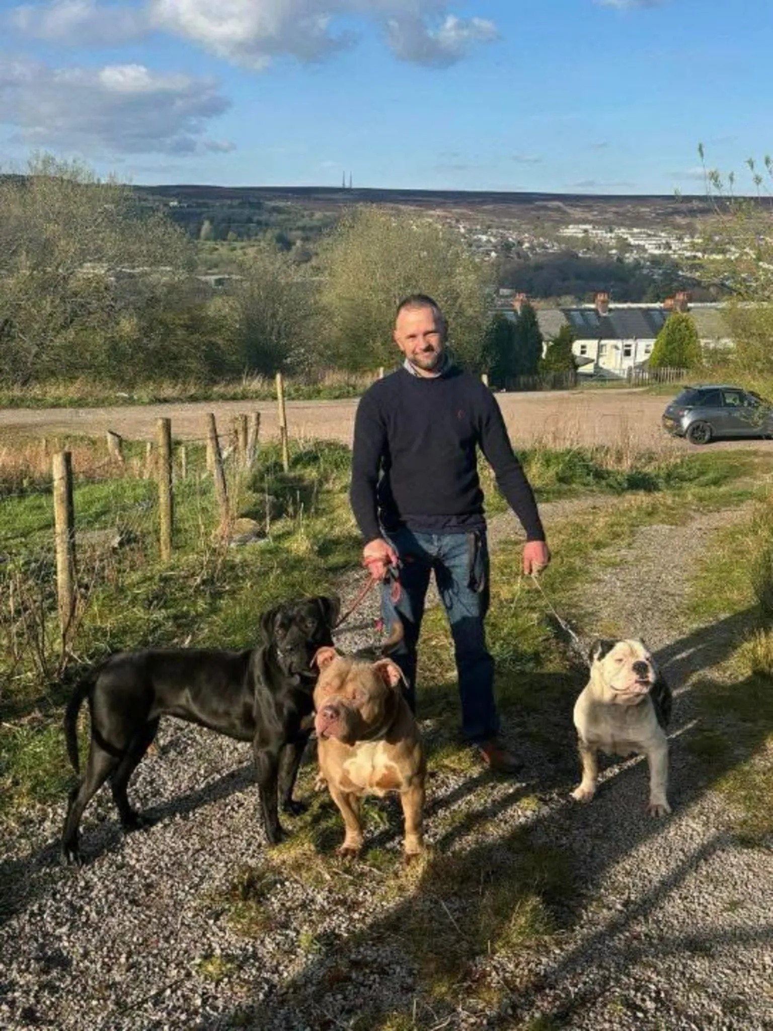Gwent Police Duane Keen wearing a dark navy jumper and blue jeans. He is standing on a track with his three bulldog-type dogs on leads and a field behind him.