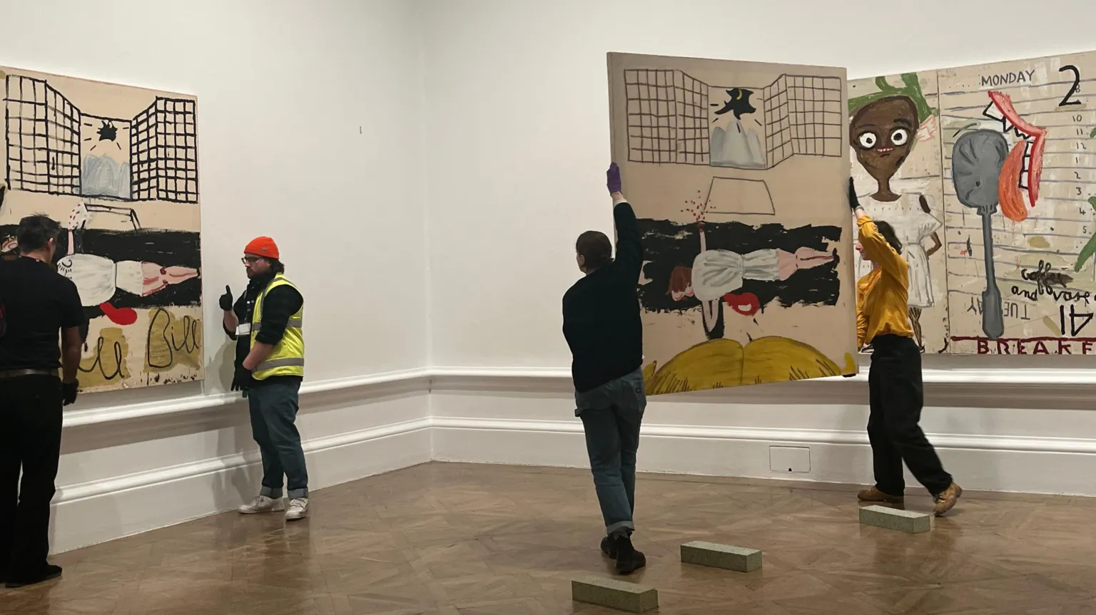 Two art handlers standing left of screen by one half of the painting Kill Bill, which partly shows a woman in white on the ground with blood on one side. T other art holders are carrying the other half of the painting towards the wall.