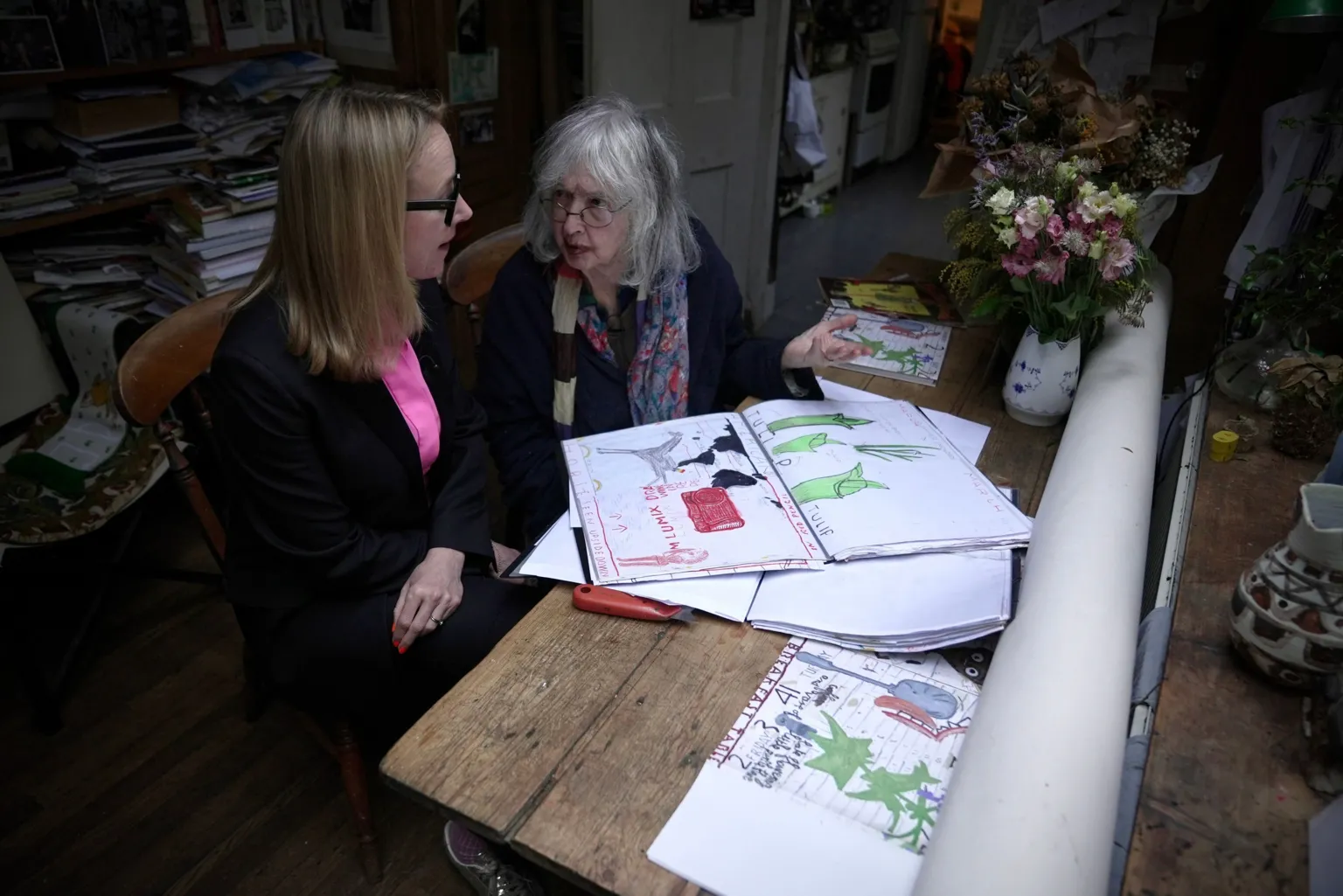 BBC/Adam Walker Katie Razzall, seated closest to the reader, in a dark jacket and pink shirt talking to Rose Wylie in a dark top and striped scarf. On the wooden table next to them are Rose's drawings - two in green, another with red, black and grey.