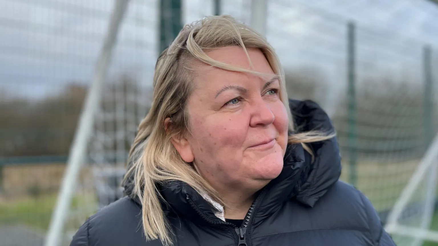 Nicola Mackenzie has shoulder-length blonde hair with some strands blown across her face. She is standing outside, next to a metal fence, and is wearing a black padded jacket. She is looking away from the camera.