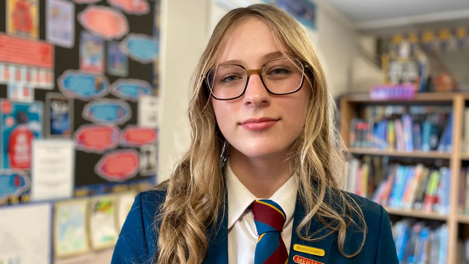 Yelyzaveta is standing in a library. She has long blond hair and is wearing glasses and a school uniform. 