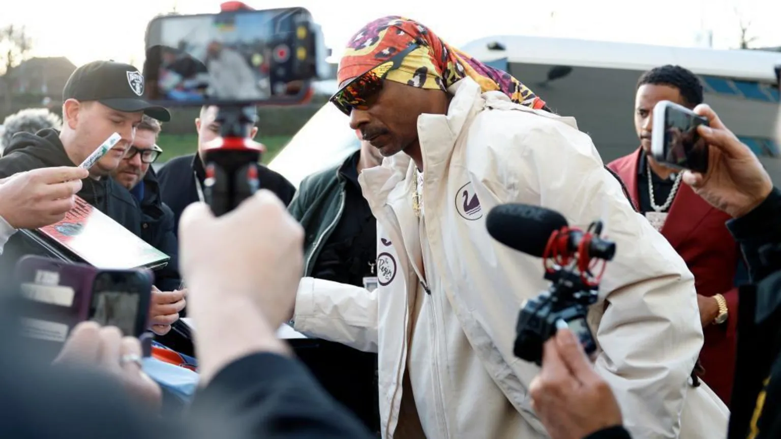  A man wearing sunglasses and a Swansea City Snoop Dog white coat is surrounded by members of the media holding phones and cameras as he arrives at the ground.