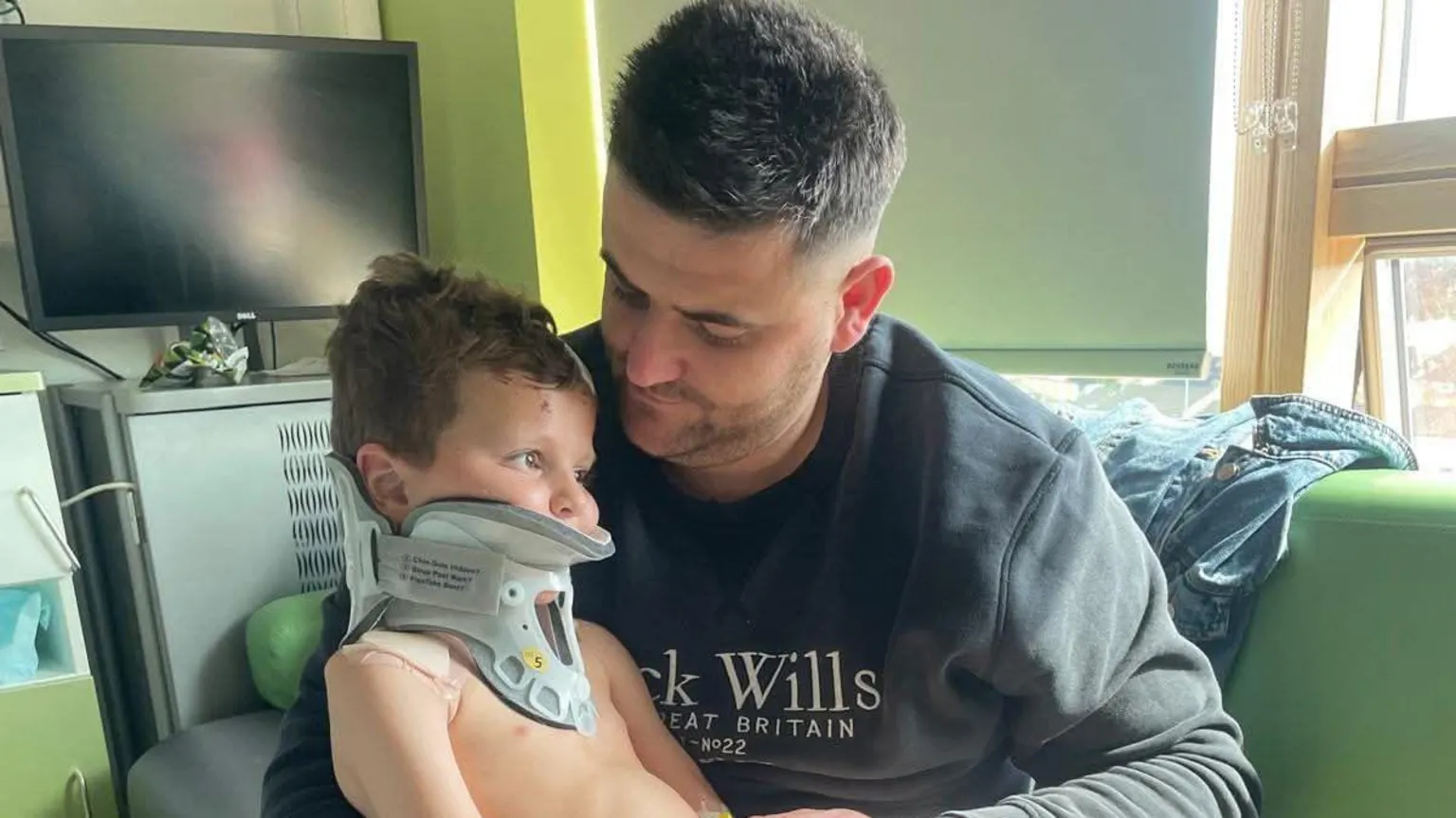Family photo A boy wearing a neck brace is sitting on his father's knee in a room in a hospital. 