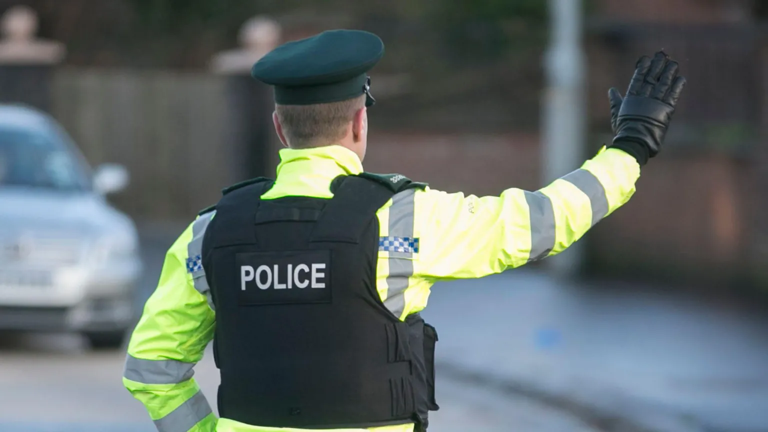  A police officer holds up their arm as they stop traffic on the street. He is pictured from behind and is wearing a police hat and vest, as well as a reflective coat. A car in the background is blurred.