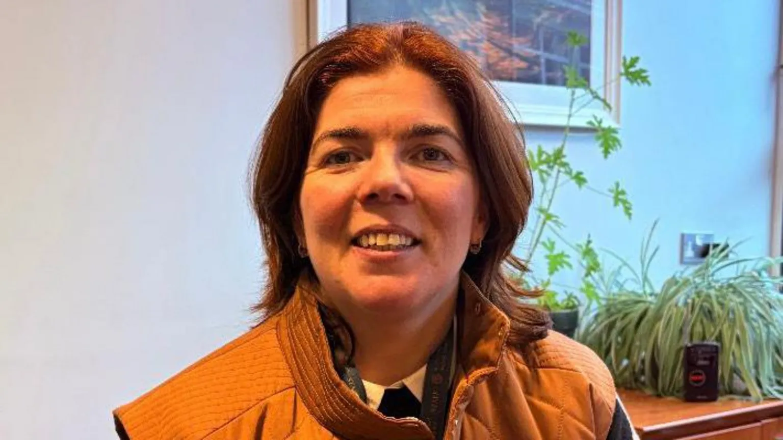 Marion McMahon A still image of Marion McMahon. She has shoulder length brown hair, an orange puffer jacket and a black jumper. She is smiling at the camera. She is sat in front of a desk with several potted plants on top of it.