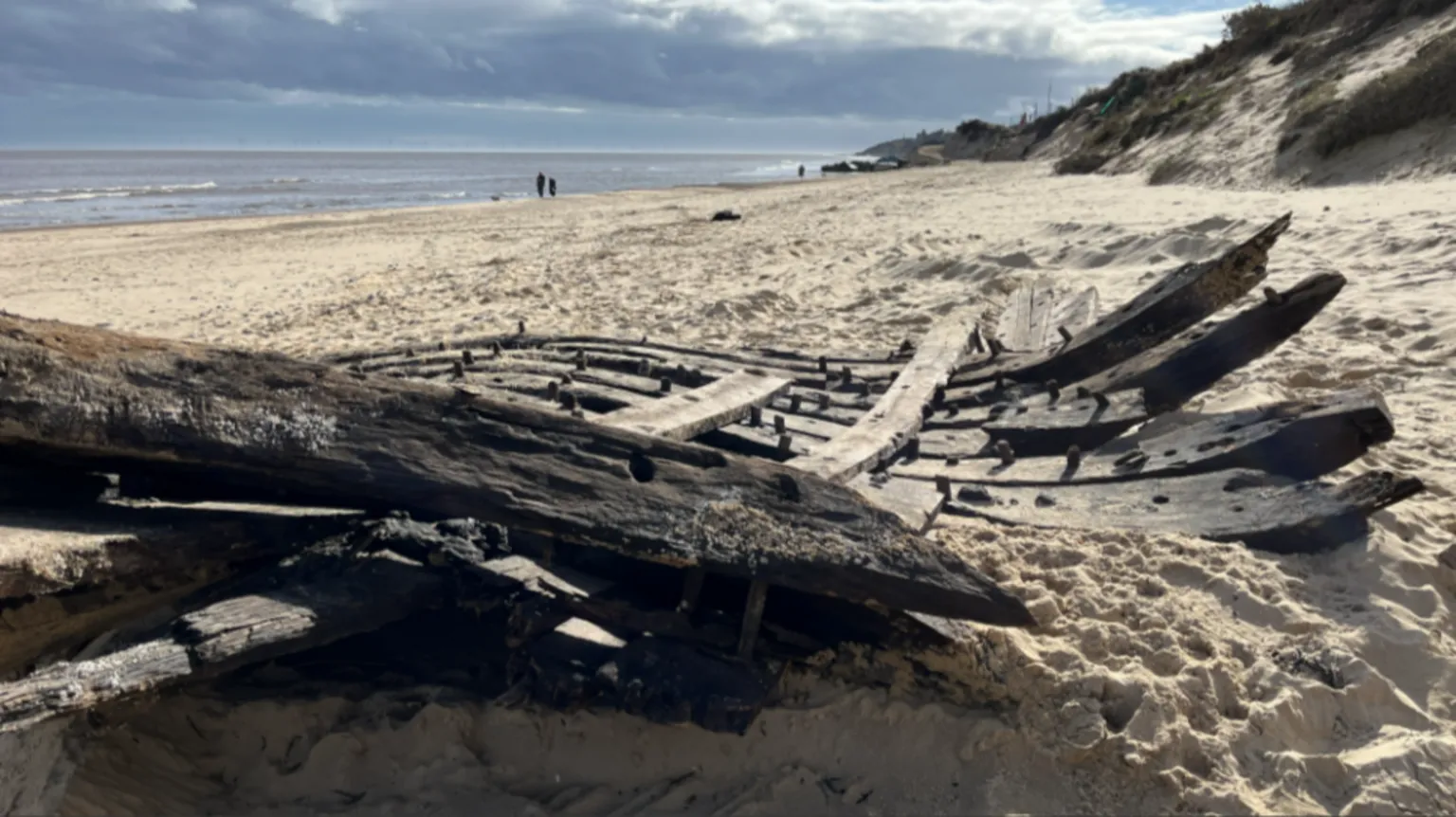 Shaun Whitmore/BBC The old timbers lie in the foreground. Beyond them, the beach - with low sandy cliffs on the right - stretches into the distance. Two tiny figures can be seen on the shoreline.