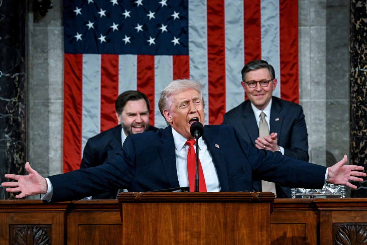 Donald Trump on stage giving a speech with both of his arms outstretched. Seated behind him are vice-president JD Vance and speaker of the house Mike Johnson.