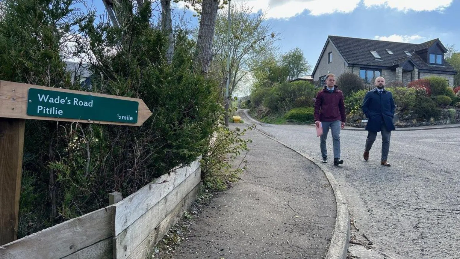 Two police detectives walk down a road with a sign saying Wade's Road, Pitilie in the foreground