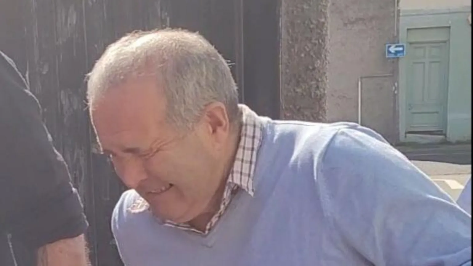 Premier News A 75-year-old man with grey hair and a blue jumper grimaces as he is led from a prison van