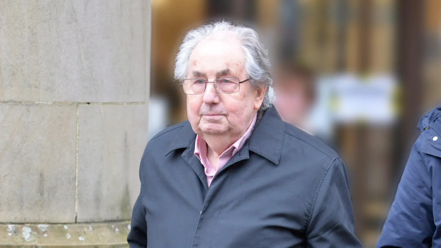 Spindrift A man in his 70s with grey hair and glasses, wearing a blue coat and pink shirt, leaves a court building