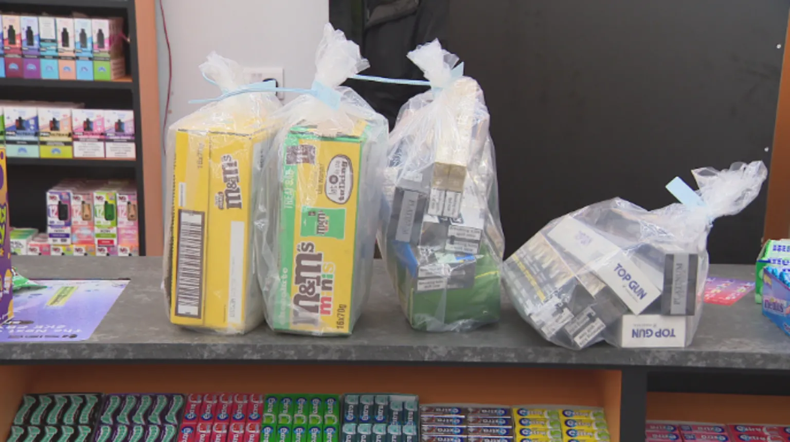 Four clear plastic bags full of illicit tobacco and cigarettes, some in M&M boxes, are sitting on a wooden counter.