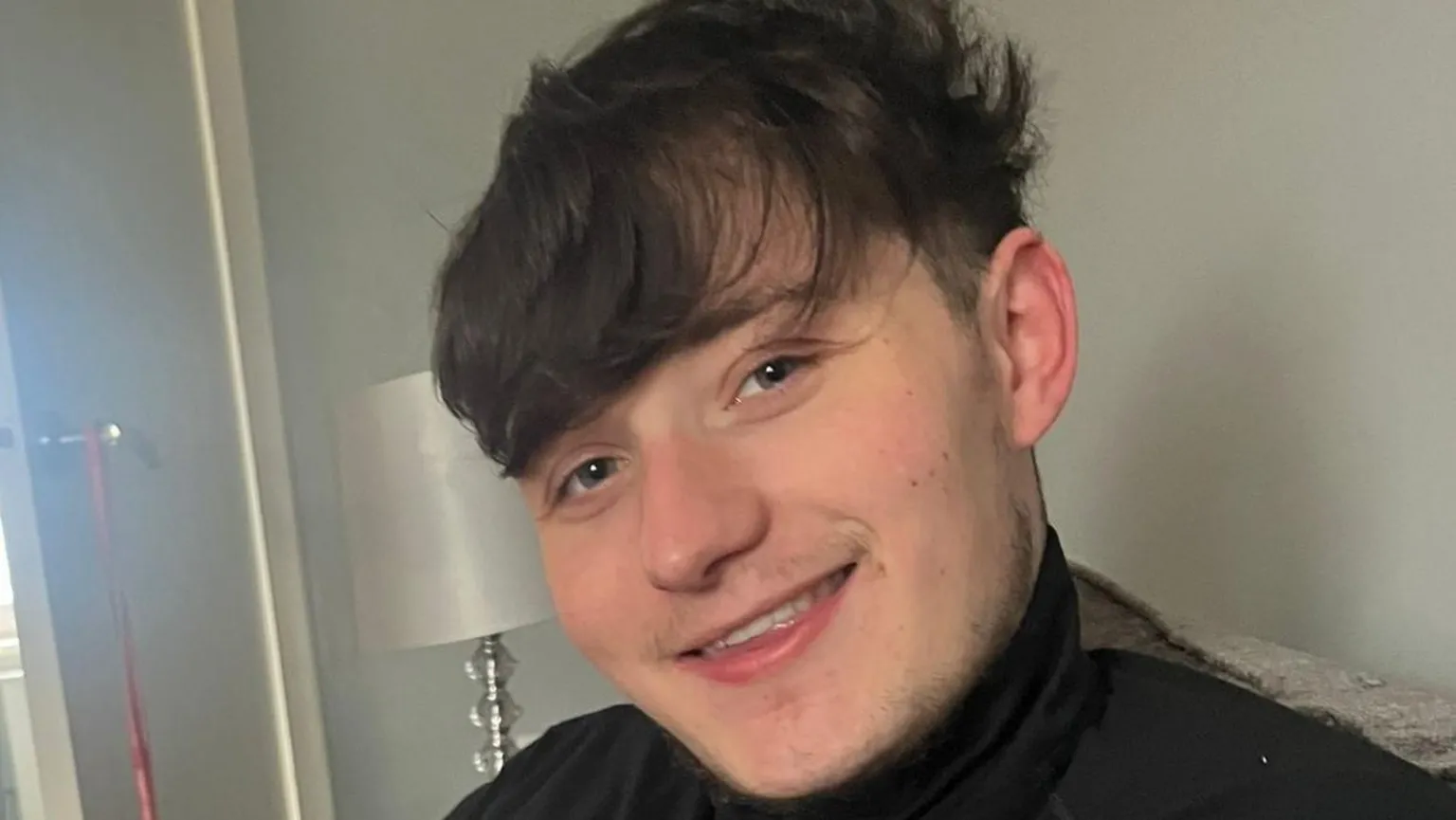 Lisa Petrie John Mcnab smiles at the camera while sitting on a couch in a living room. He has short dark brown hair and wears a dark top.