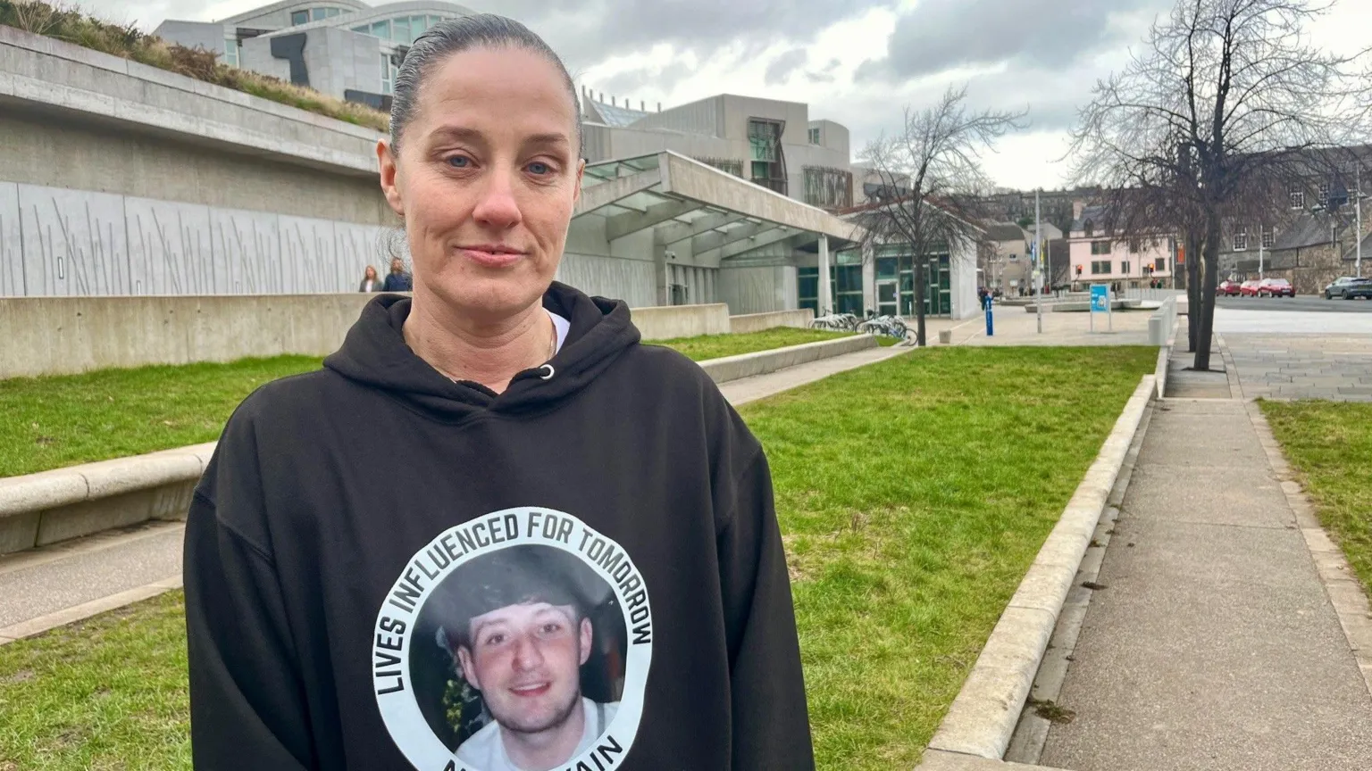 Lisa Petrie standing outdoors near a modern concrete building and grassy area, wearing a black hoodie with a circular graphic and text on the front. Pathways, trees without leaves, and cloudy skies are visible in the background