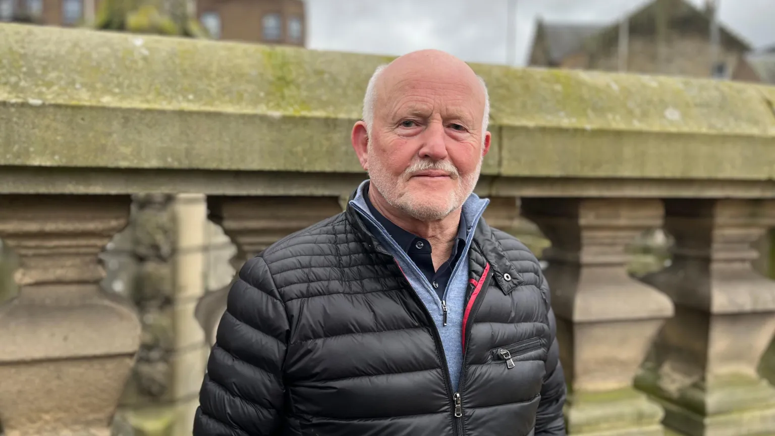 A bald man with grey beard, wearing a black jacket, looks at the camera