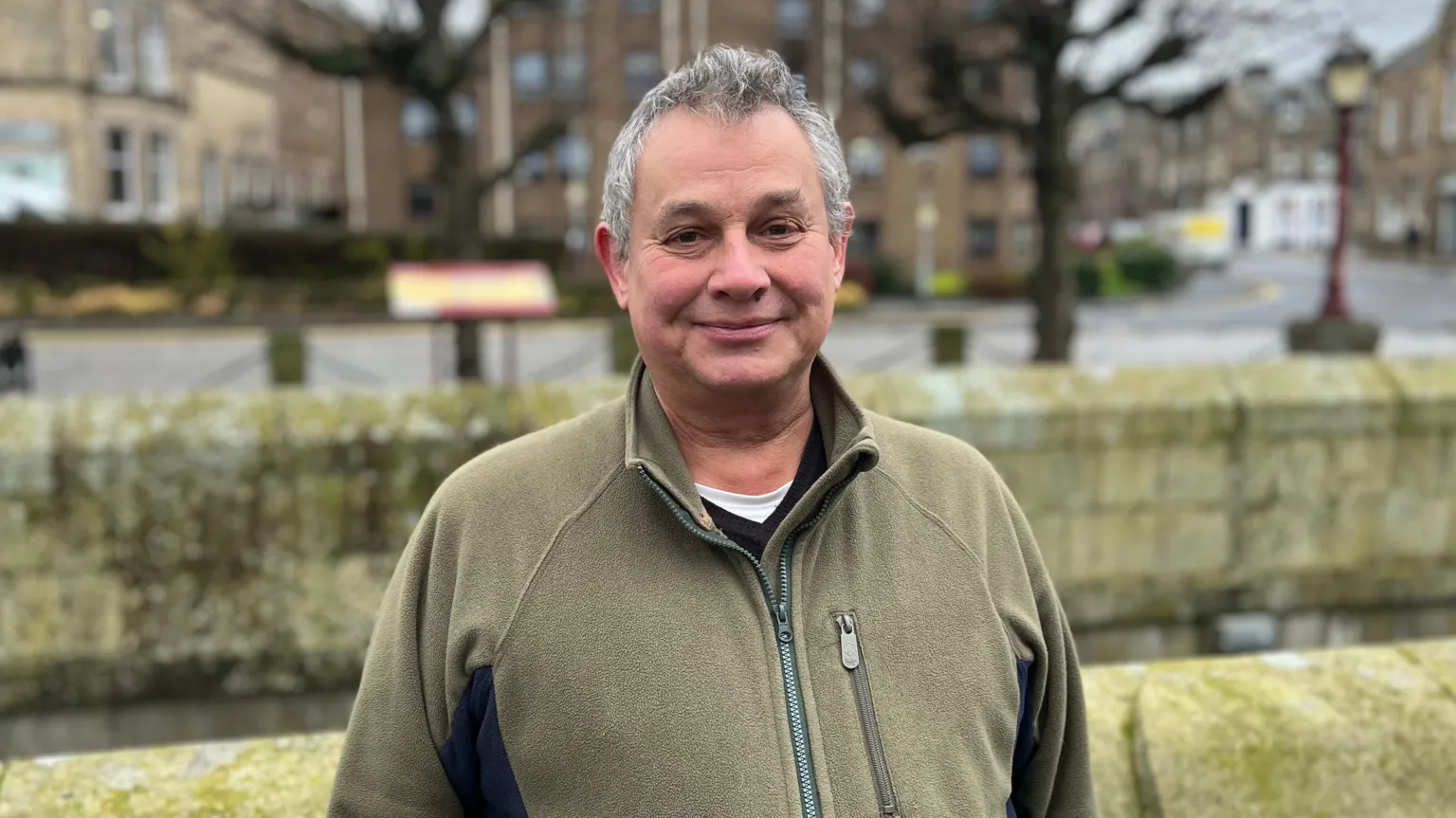 A grey haired man with a light green jacket looks at the camera, with trees and stonework in the background